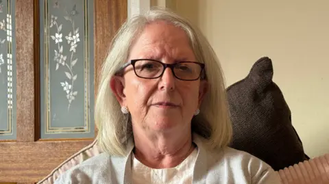 Audrey Buckham, wearing black rimmed glasses, a white shirt and a light grey jacket, has blonde hair and two pearl-appearing earrings and is sitting on a sofa inside her living room.