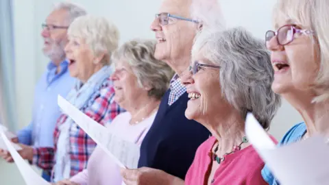 A group of six elderly people - four women and two men - standing in a line and singing from a choir lyrics sheet. They are standing in a brightly lit room, smiling and looking off to the left of the camera.