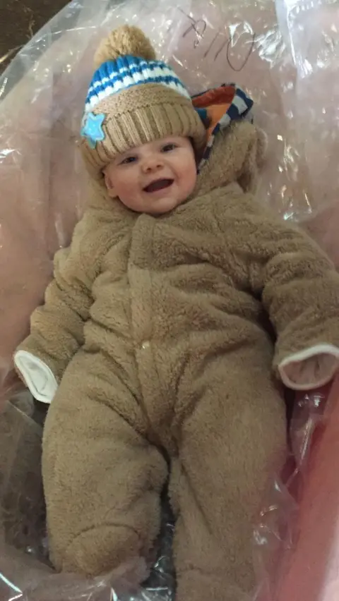 Family handout A very young baby is smiling in a pram while wearing a thick winter pram suit. The baby has a wooly hat on his head. 