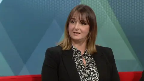 Ms Owen has cropped hair and a fringe. She is wearing business attire and sitting on a sofa in a television studio. 