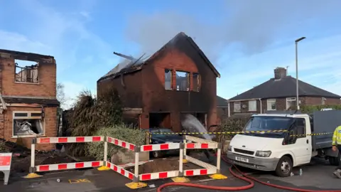 A house that has been destroyed by a fire. All that remains is a shell of brick work, charred timber, rubble and ash.