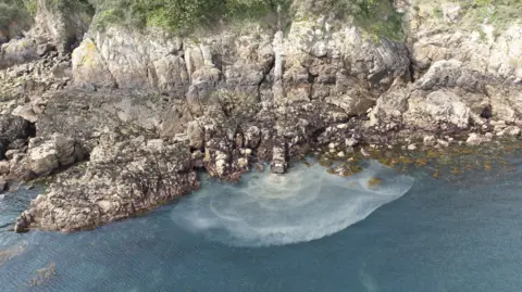 A drone image of the cliff face at Soldier's Bay. It shows the outfall pipe, covered in brown granite. Emerging from that into the sea is brown water, which is seeping into the blue sea. 