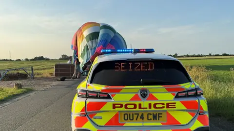 Cambridgeshire Police Brightly coloured deflated hot air balloon on side of rural road near fields. A police car is in front of the balloon.