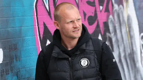 Liam Manning a man who is standing outside in front of a wall covered in graffiti. He is looking away from the camera and is wearing a black jacket and black gilet. 