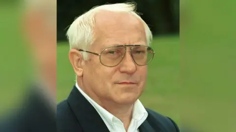 BBC Oleg Gordievsky looks at the camera, has short white hair and is wearing a white shirt under a dark suit jacket. There is a lawn in the background.