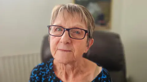 Enid has short light brown hair and is wearing dark rimmed narrow glasses. She is looking directly at the camera and is wearing a blue and black patterned top. 