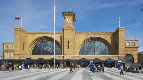 Getty Images Outside King's Cross railway station in London, showing a yellow brick-built building, with two very large glazed arches into the station, a tower in the middle with a clock, and people walking in front of the station on a grey striped public square