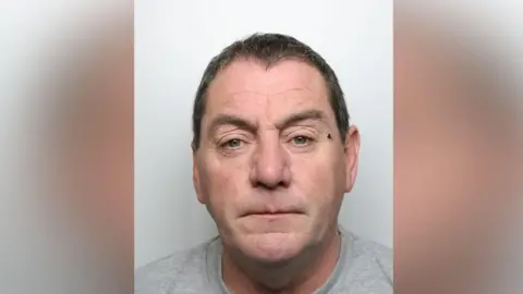 A mugshot of a man in his fifties. He had dark short hair and what appears to be a small cut close to his left eye. He is staring directly into the camera.