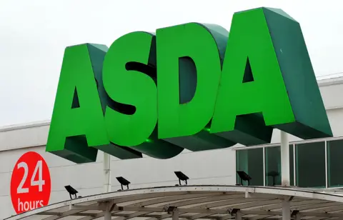 The large Asda sign, in giant green lettering, is fixed at the entrance of an Asda store. It is a cloudy day.