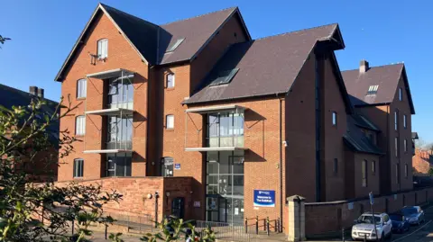 A red-bricked five storey building with various gabled roofs and glass balconies. Cars are parked along the right-hand side of the building and a blue sign is on the wall by the entrance that reads "Welcome to The Guildhall".