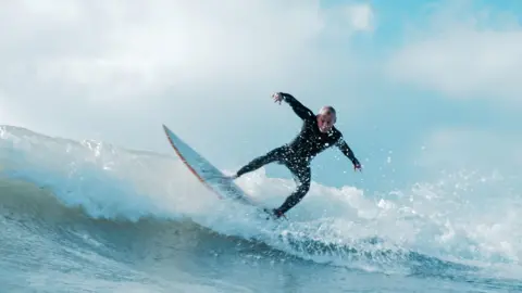 Fine Rolling Media Mark standing on his board in a wave of breaking white water. Mark is leaning over to the right