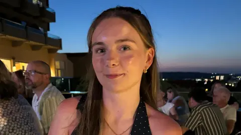Caitlin Graham A close up photo of Caitlin, a young woman with long brown hair. She is smiling against a dusky night sky.