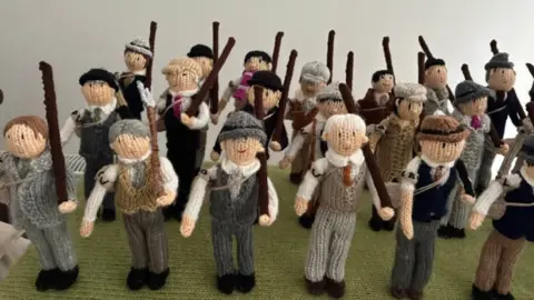 Rheged Several knitted soldiers holding shotguns, some wearing waistcoats and hats. They are standing in three rows, as if on parade.