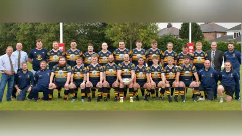 A team of male rugby players wearing a blue and yellow kit consisting of a short sleeved rugby top, shorts and socks. There are two men with light shirts, patterned yellow ties and jeans, one person with a light shirt, stripy yellow tie and grey suit, two men with blue sports tops and bottoms and one lady with a blue sports top and dark shorts. They are on a grass pitch with goal posts behind. There are some trees and houses.