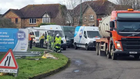 Chris Jones Four transit vans and a tanker, with several workers in hi vis and hardhats, standing beside a fenced off section of road.