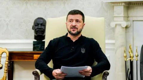 EPA Zelensky wearing a black long-sleeved t-shirt and holding some A4 paper, has a frowning expression as he sits in a white armchair in the White House next to Biden at a news conference. Behind him is a fireplace and cast iron bust