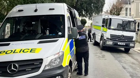 A police van and a police officer stood next to it. To the right is a bomb disposal truck. There is take in the background. 