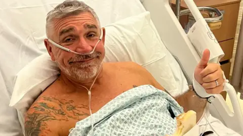 A man with greying hair lies in a hospital bed in a hospital gown. He is giving a thumbs up to the camera.