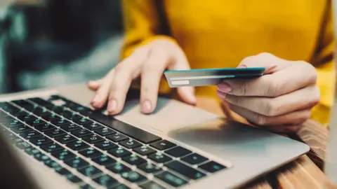Getty Images A person sits at a laptop computer. We can see a close-up of their hands. They are holding a credit card, and it appears that they are typing its numbers into the computer.