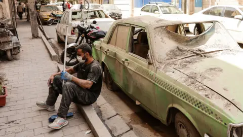 A man with a mask over his nose and mouth and gloves on his hands sits on the pavement holding a broom while he takes a break from clearing debris, while behind him stands a blown-out green taxi with shattered windows and dents in its side, and other vehicles behind it lining the street. Taken in Tehran on Thursday.