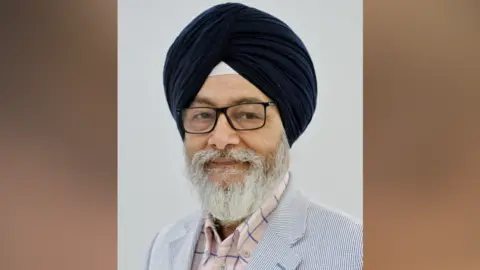 Slough Borough Council Councillor Manku is smiling at the camera and has a dark turban and has a white beard. He is wearing a light blue suit and pink and blue open-necked shirt. 