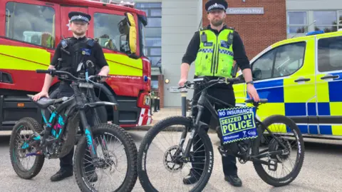 Two police officers stand by a fire engine and police car, each with an e-bike in front of them. One bike has a poster on it that says "No insurance. Seized by police". The officers are stood outside Cheshire Constabulary's building.