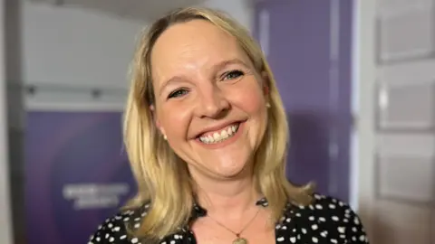 Kate Wright smiles at the camera. She has shoulder length blonde hair. BBC Jersey branding is visible in the background.