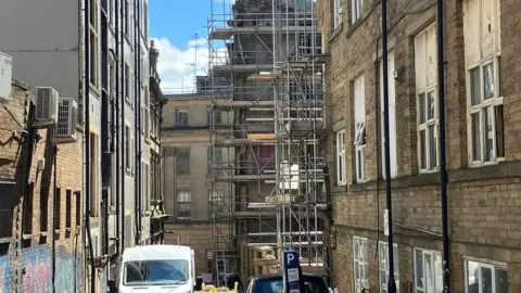 A view of a street with tall brick buildings and a building surrounded by metal scaffolding at the end of the road.