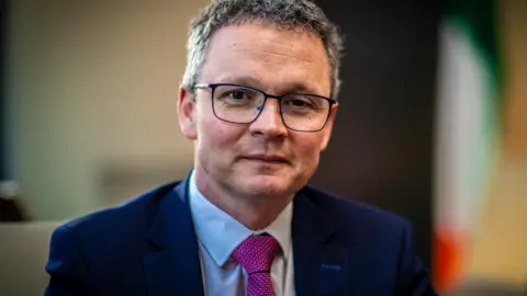A man wearing a dark blue suit jacket, white shirt, and bright pink patterned tie, sitting indoors with soft, warm lighting and a partially visible tricolour flag in the background.