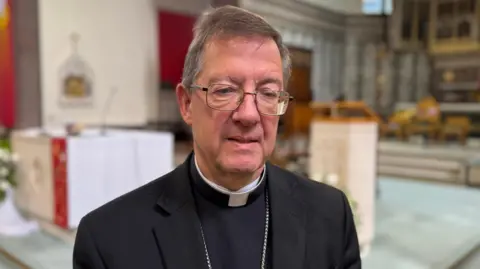 John Sherrington wears a black suit jacket and top, with a white dog collar and silver-rimmed glasses. He has grey hair and stands in a cathedral, which is blurred out in the background.