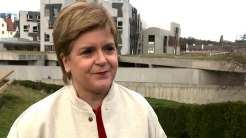 Nicola Sturgeon stands in front of the Scottish Parliament, she has short hair, wears a red top and cream jacket