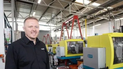 Clarity Plastics Capul și umerii unui bărbat, zâmbitor, purtând o cămașă neagră, stând într-o fabrică cu un rând de unități de producție galbene în fundal