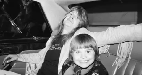 Getty Images A black and white picture of a man with long hair, a fringed jacket and dark t-shirt with a smiling younger child both in the back of a car.