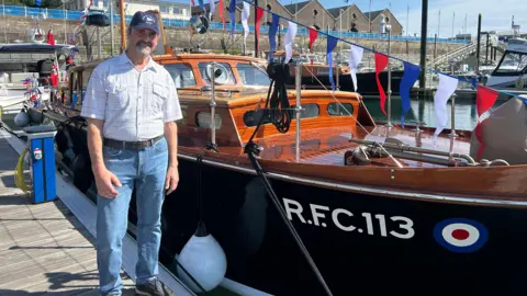A man called Guy Riviere stands next to a wooden boat called RFC 113. Riviere is wearing a baseball cap, short-sleeve white chequered shirt and blue jeans. Red, white and blue bunting is on the boat.