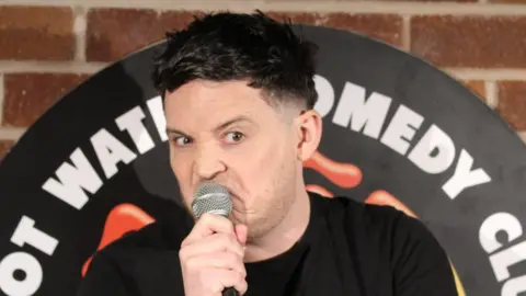 Des Hinks Des Hinks talking into a microphone while on stage at a comedy club. He has short black hair and is wearing a black T-shirt.