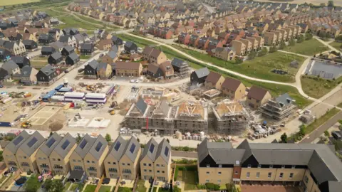 An aerial image of Northstowe showing pockets of new and occupied homes and green spaces all surrounding a central area which is still under construction with scaffolding, diggers, piles of building supplies.