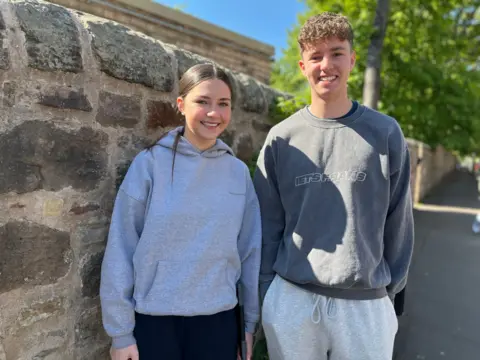 A girl in a grey hoody and a boy in a grey sweatshirt are standing by a grey wall, smiling.
