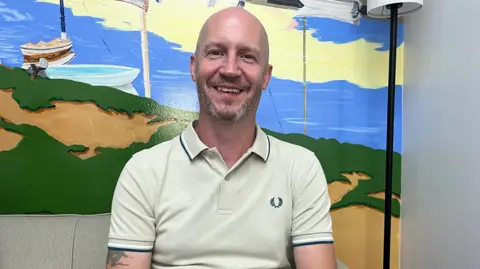 David Shaw sits in front of a mural depicting the Norfolk coast in yellow, green and blue. He is wearing a cream polo shirt. He is bald and has grey stubble. 
