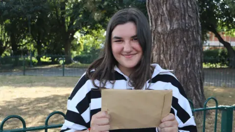 Barnardos A picture of a girl with brown hair and a black and white jumper. She is holding a brown envelope in her hands.