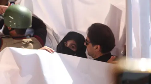 EPA Bushra Bibi and Imran Khan were protected by a white sheet as they arrived at the court. Bibi has turned to look at the camera, her hair is covered as well as her nose and mouth.