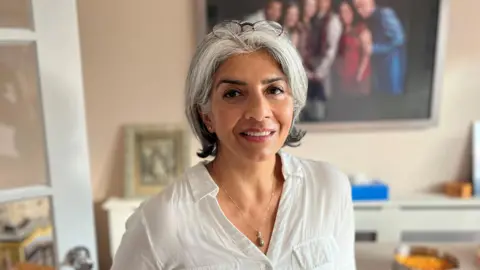 BBC Pragna Hay wearing a white top, with shoulder-length grey hair. She has reading glasses perched on her head. She is wearing a necklace with a pearl pendant on it. She is standing in her lounge and behind her are a couple of family photos.