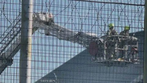 Jamie Niblock/BBC Firefighters in the cage of an aerial ladder platform. There appear to be two of them. They are approaching a roof. The shot has been taken through a wire fence at Highpoint Prison