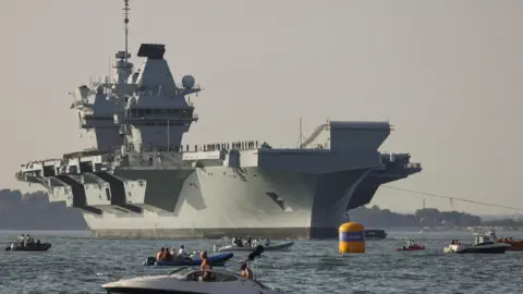 Royal Navy Queen elizabeth on the water, with lots of smaller boats in front of it. The sky is orange.