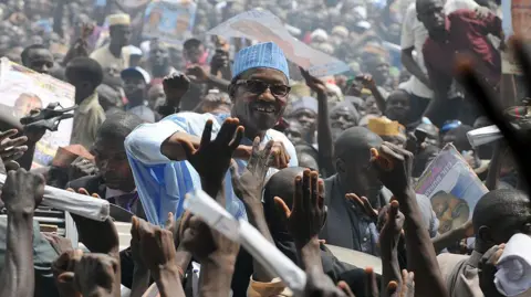 AFP via Getty Images At a packed rally, supporters clamour to touch Buhari, who stands above the crowd, smiling. He wears a blue top and blue hat.