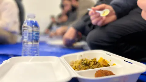 Jon Wright/BBC A close up of a polystyrene takeaway container with rice and samosas inside. People are in the background sitting on the floor and eating.