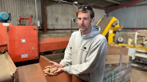 Luke Abblitt is wearing sunglasses on his head, a grey hoodie and has short dark brown hair . He is standing in a corrugated iron building, surrounded by machinery and is holding a handful of potatoes. 