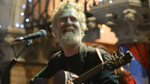 Nur Photo/Getty Images Glen Hansard sonríe mientras toca la guitarra a las afueras de un teatro de Dublín en la noche de Nochebuena de 2019. Es un hombre con cabello pelirrojo canoso y barba a juego. Lleva un blazer verde oscuro sobre una camiseta oscura.