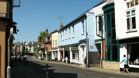 Geograph A quiet street lined with pastel-coloured shopfronts and mixed architectural styles, viewed on a sunny day. A few pedestrians walk along pavements beneath a clear blue sky, with long shadows stretching across the road.