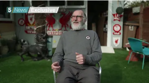 Stefan Dettlaf has white hair and beard and is wearing a grey sweatshirt and trousers. He sits on a garden chair in front of the rear of his house which has Liverpool FC favours on the wall.