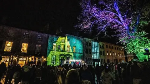 Robin Zahler A light projection  in green colours on buildings on Dalton Square in the dark, with a lit up tree to the right and lots of people milling about in front of the buildings